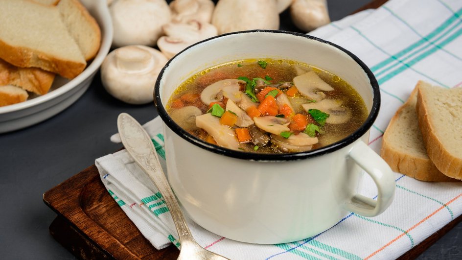 Наваристый суп с курицей, шампиньонами и клецками в тарелке на деревянной подставке рядом с хлебом и ложкой