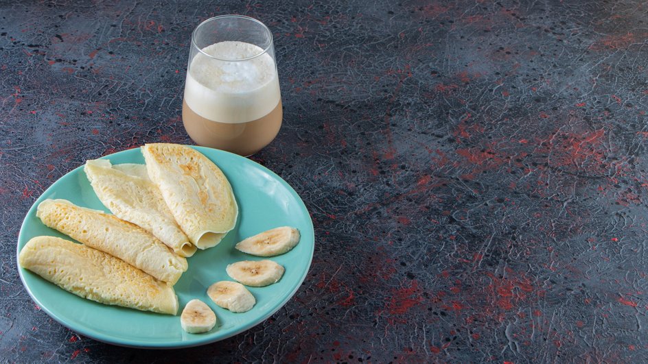 Аппетитные блины без муки на молоке лежат на тарелке рядом с кусочками банана и стаканом какао Аппетитные блины без муки на молоке лежат на тарелке рядом с кусочками банана и стаканом какао