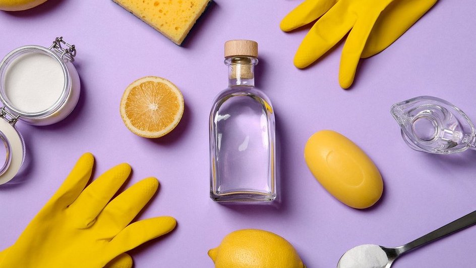 Бутылка с уксусом лежит на фиолетовом столе рядом с резиновыми перчатками, губкой для посуды, лимоном и ложкой
