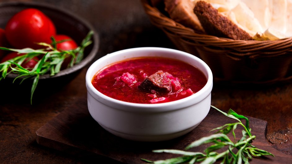 Борщ без капусты с курицей в супнице стоит на деревянной доске рядом с зеленью, чашей помидоров и корзинкой хлеба на столе Борщ без капусты с курицей в супнице стоит на деревянной доске рядом с зеленью, чашей помидоров и корзинкой хлеба на столе